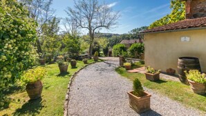 Un camí de grava que condueix a un edifici rústic amb teulada de rajola, envoltat de plantes en testos i arbres.