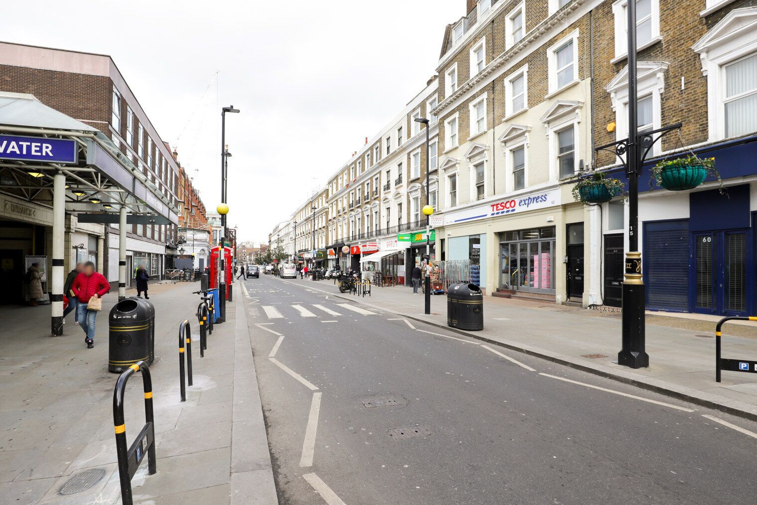 Rua com lojas, incluindo uma Tesco Express e um bebedouro.