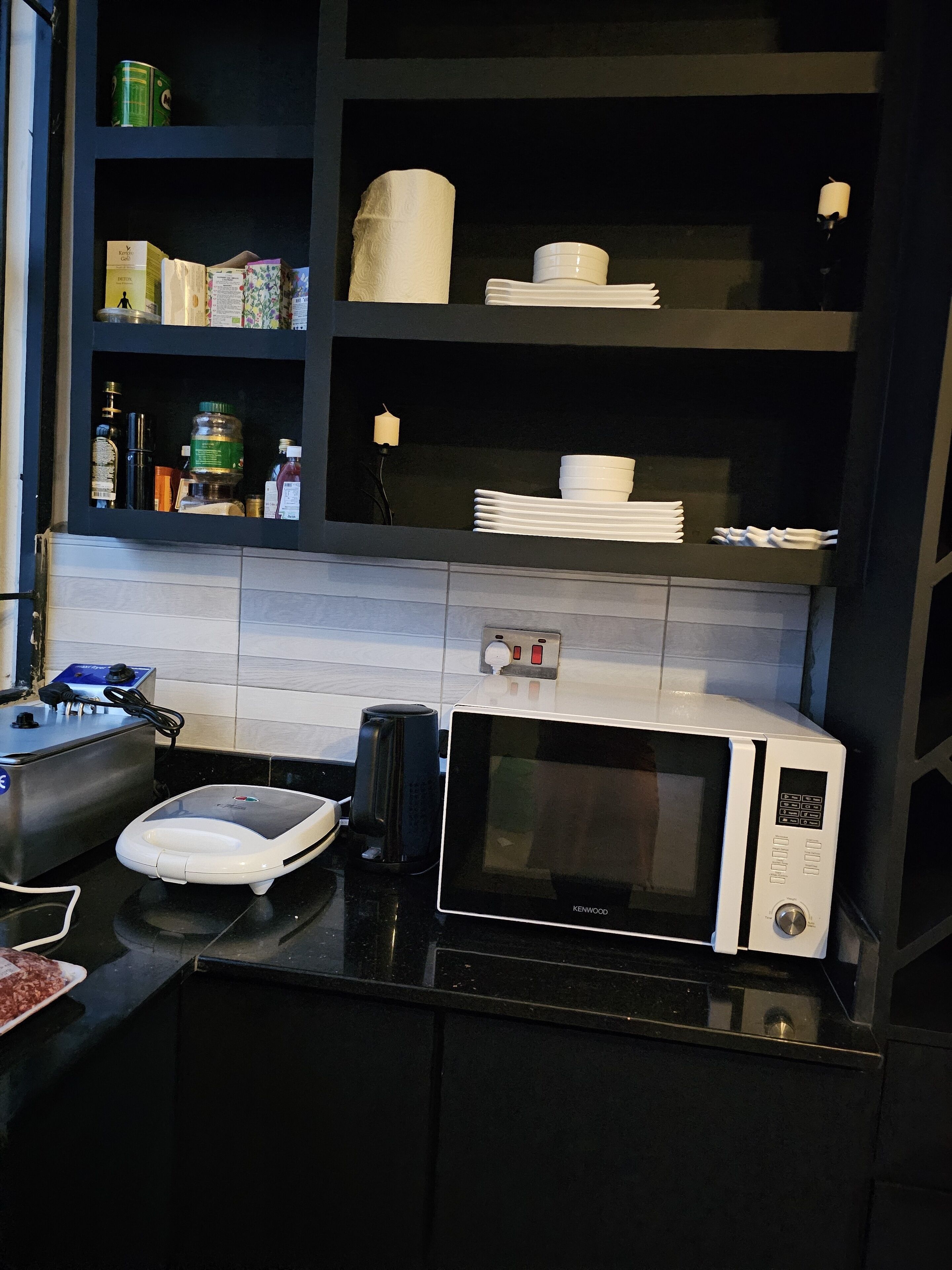 A modern kitchen with a microwave, coffee maker, and a scale. There are shelves with various items including a can, a box, and a bottle.