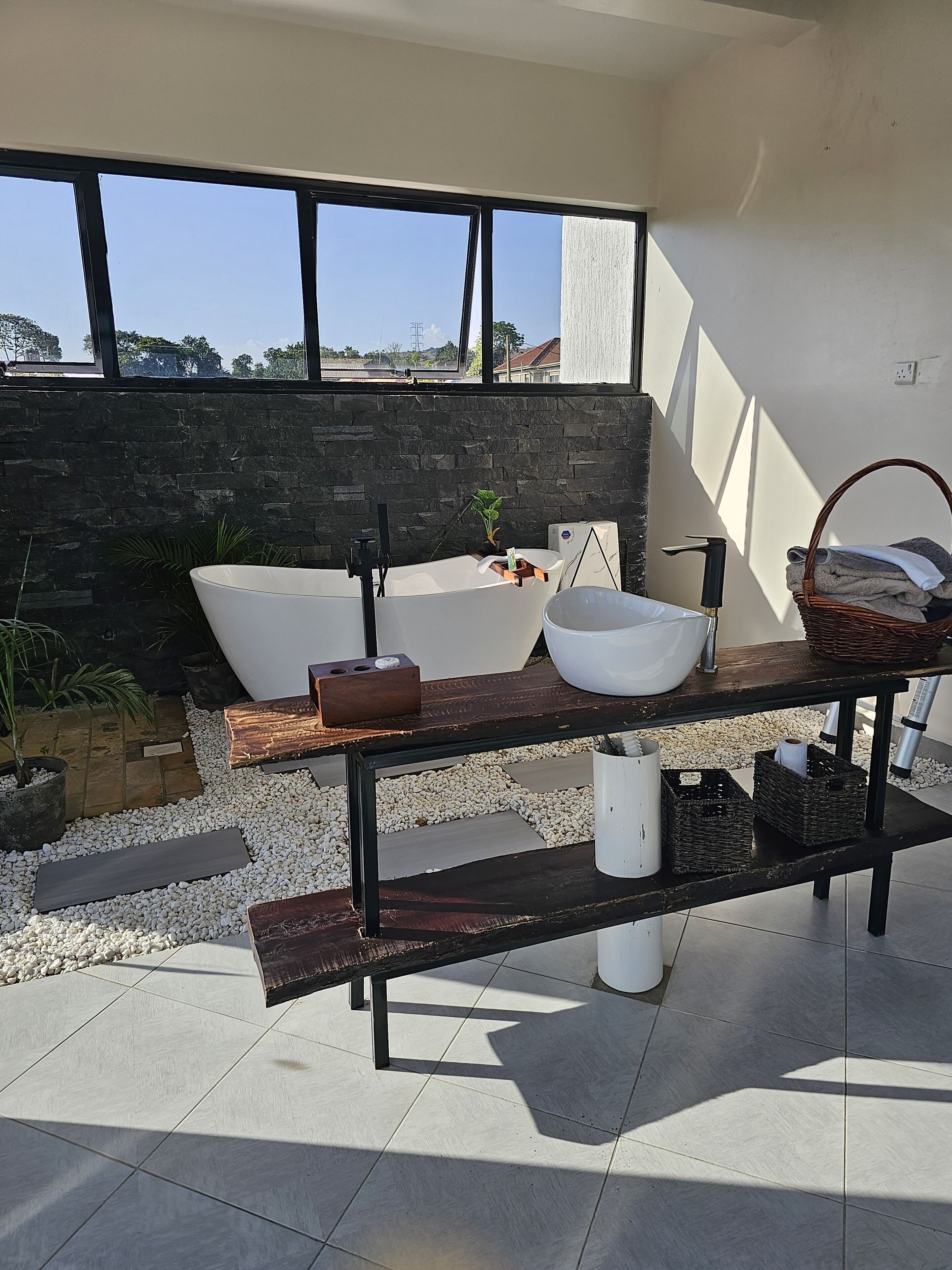 Un baño exterior moderno con una bañera independiente, un lavabo y una amplia pileta. Hay una mesa de madera con una cesta y toallas, así como una planta en maceta. El espacio está pavimentado con baldosas y rodeado de guijarros.