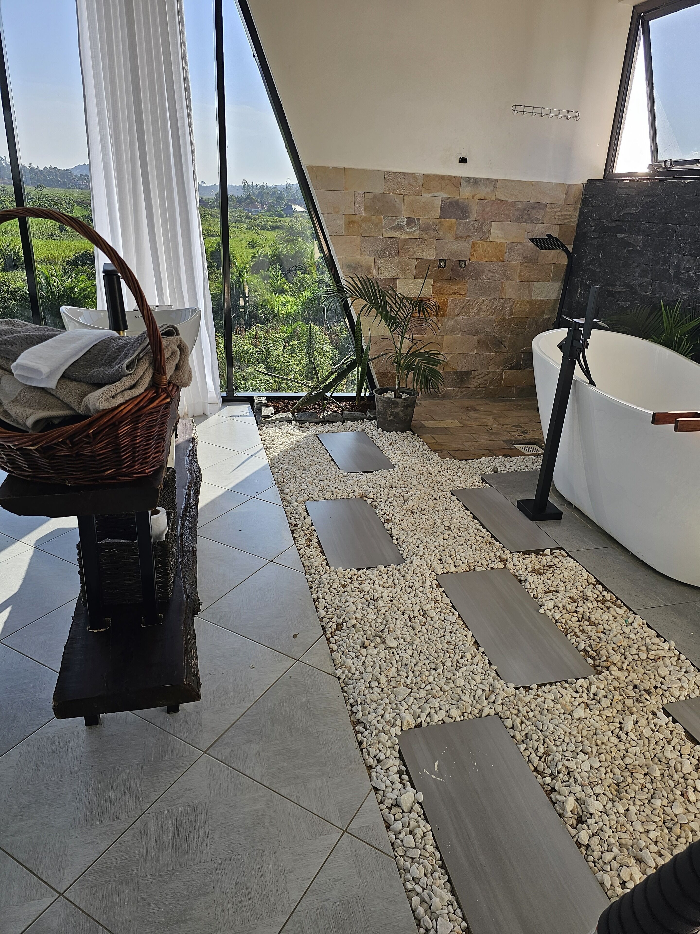 A modern bathroom with a stone wall, a white bathtub, and a large window overlooking greenery.