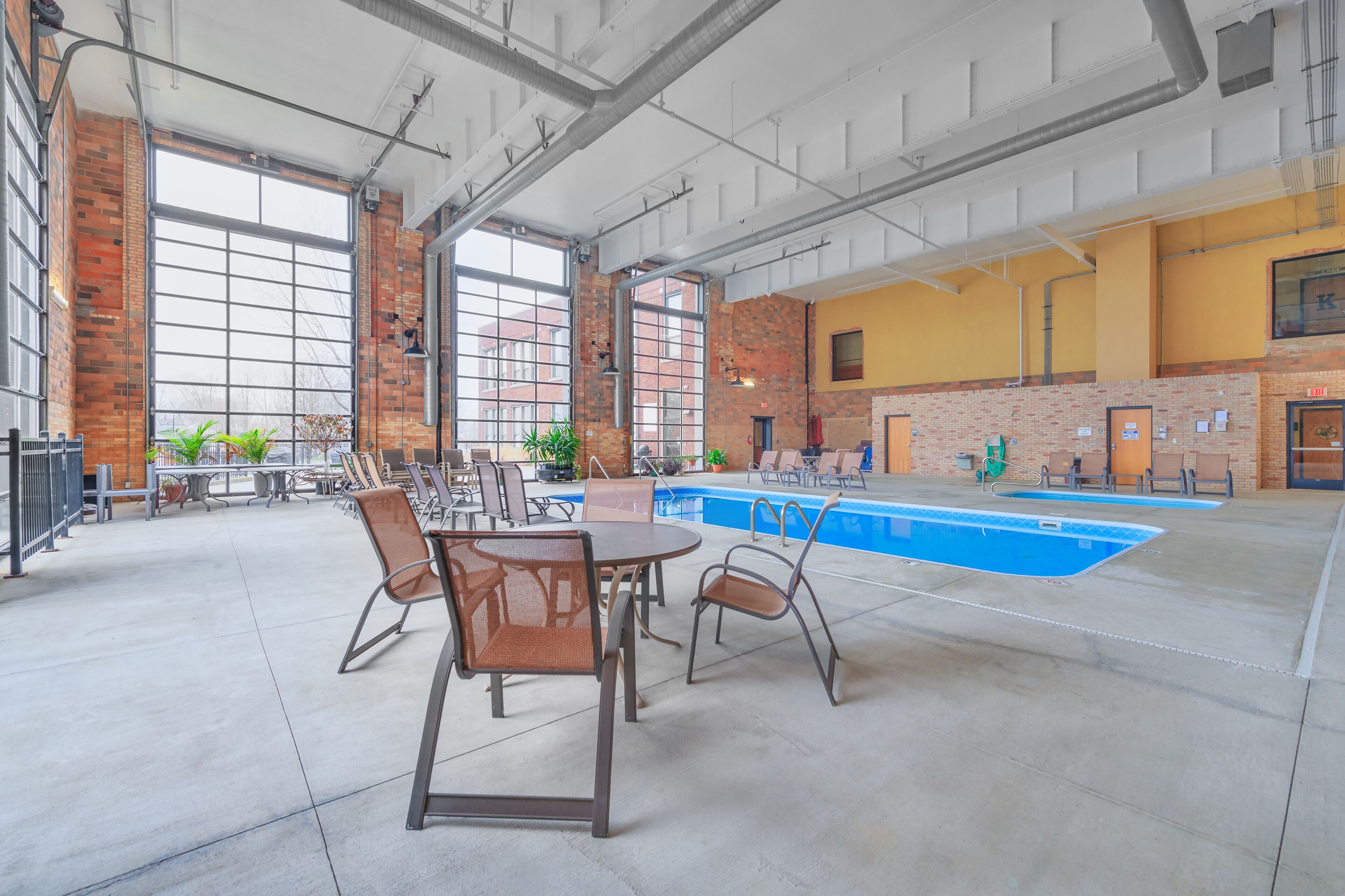 An indoor swimming pool area with a brick wall, large windows, and seating around the pool.