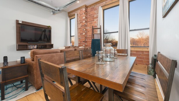 Een eetgedeelte met een houten tafel en stoelen, een aan de muur bevestigde televisie en een bakstenen muur met grote ramen.