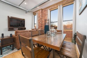 Un comedor con mesa y sillas de madera, un televisor montado en la pared y una pared de ladrillo con ventanas grandes.