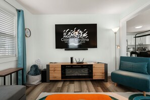 A modern living room with a flat-screen TV mounted on the wall, a wooden entertainment unit, a teal sofa, and a kitchen area visible through an open door. - Good Vibes Soundside: Cute, Soundside Home, Short Stays anytime with Hot Tub! (Kill Devil Hills)