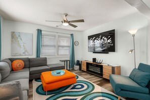 Un salon moderne avec un canapé gris, un fauteuil turquoise et un pouf orange. Un téléviseur à écran plat est fixé au mur, et un ventilateur de plafond est visible.