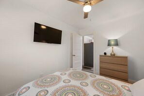 Un dormitorio con cama, televisor montado en la pared, ventilador de techo, cómoda y una puerta que da a un armario.
