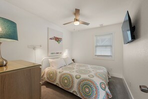 Une chambre à coucher comprenant un lit, un ventilateur de plafond, un téléviseur fixé au mur, une fenêtre avec des stores et une carte géographique accrochée au mur.