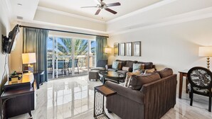 A well-lit living room with a sofa, coffee table, and a ceiling fan. A balcony with curtains is visible through the glass doors.