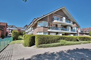 Un immeuble résidentiel à plusieurs étages, doté de balcons, d’une allée pavée et de haies bien entretenues.