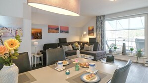 A modern living room with a large sofa, a dining table with plates of food, and a large window with curtains. - Seesternresidenz Appartement 21 - Seesternresidenz (Scharbeutz)