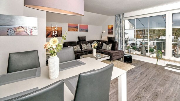 A modern living room with a sofa, coffee table, and dining area. There are large windows with a view of the outside. - Seesternresidenz Appartement 21 - Seesternresidenz (Scharbeutz)