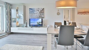 A modern living room with a flat-screen TV, a dining area with a table and chairs, a large window with curtains, and a bookshelf. - Seesternresidenz Appartement 21 - Seesternresidenz (Scharbeutz)