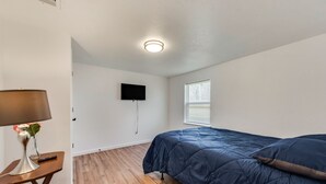 A bedroom with a wooden floor, a blue bedspread, a mounted TV, and a lamp on a small table. - Close to main attractions and Down Town Fort worth (River Oaks)