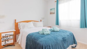 A hotel room with a wooden bed, white bedding, a blue blanket, and a small table with a lamp.