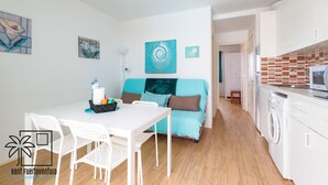 A modern kitchen with a microwave, sink, and washing machine. There is a dining area with a table and chairs, and a sofa with cushions.