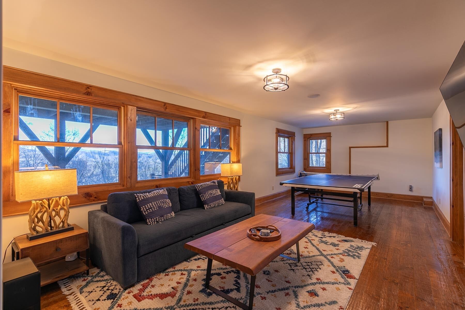 A spacious living area with a sofa, coffee table, and a ping pong table. Large windows provide a view of the outdoors.