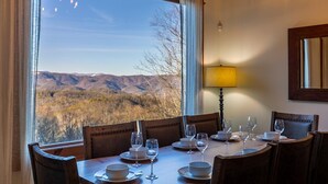 Una mesa de comedor dispuesta con platos, vasos y una lámpara, con vistas a la montaña a través de amplias ventanas.