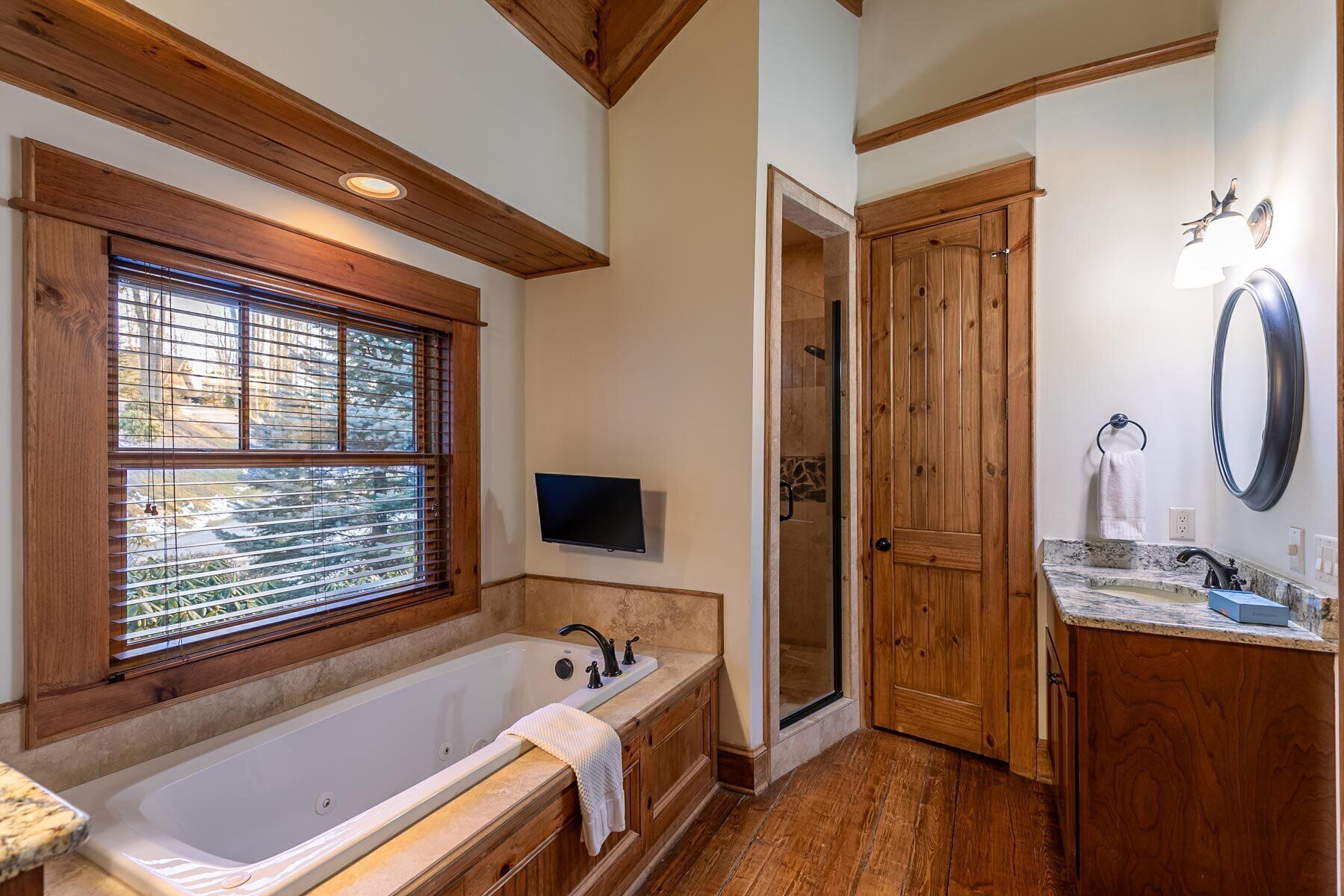 Une salle de bain avec une baignoire, un meuble-lavabo en bois, une douche et une grande fenêtre avec des stores.