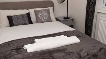A neatly made bed with a dark grey blanket and white towels, a framed picture on the wall, a bedside lamp, and a window with curtains.