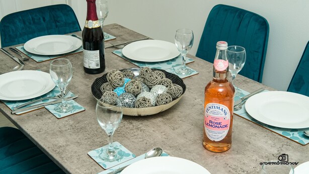 Una mesa de comedor dispuesta con platos, cubiertos y vasos, una botella de limonada de rosa, y una cesta de bolas decorativas.
