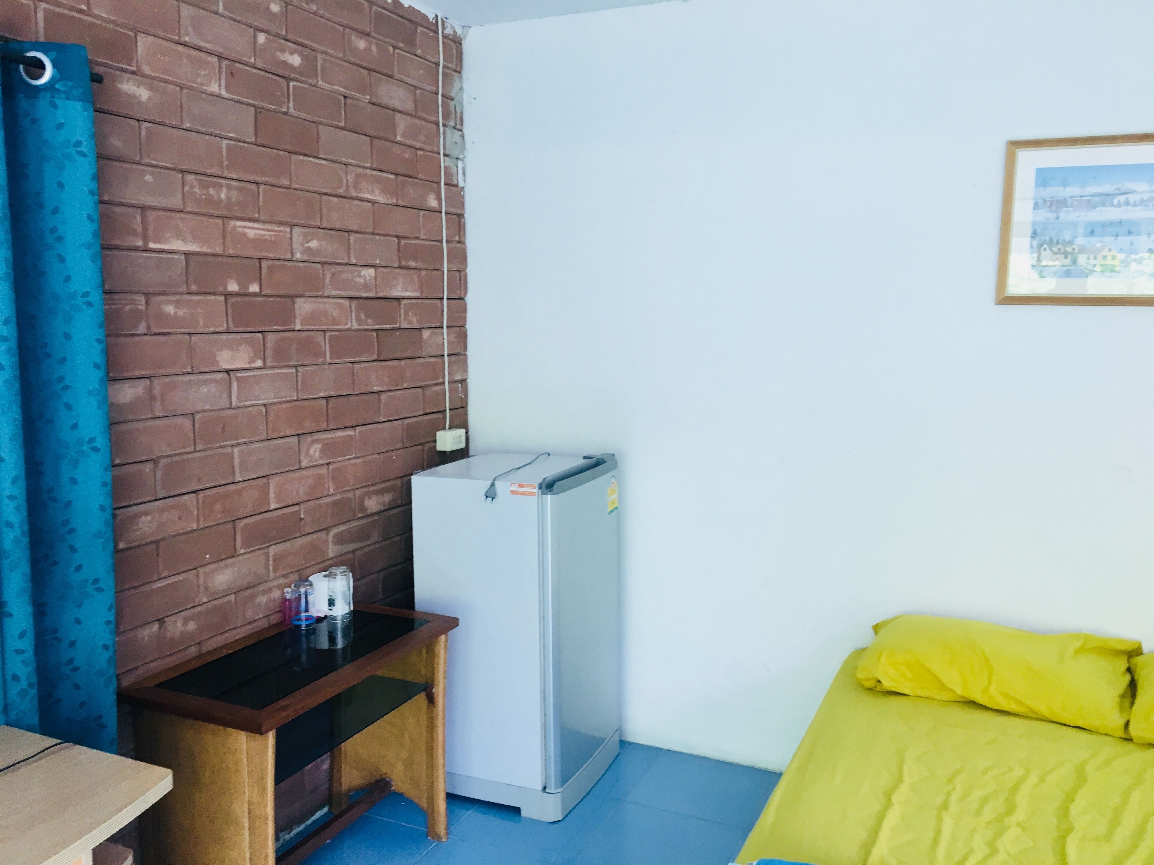 A room with a brick wall, a small table with a refrigerator, a bed with yellow bedding, and a framed picture on the wall.