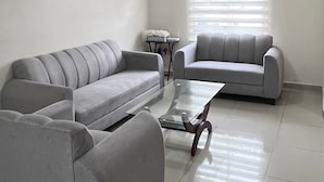 Ein modernes Wohnzimmer mit einem grauen Ecksofa, einem Glastisch und einem Fenster mit Jalousien.