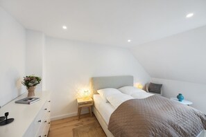 A bedroom with a bed, bedside tables, a nightstand, a bedside lamp, a vase with flowers, and a small table with a book and a cup. - Lykke - Hochwertige Modern-skandinavische Ferienwohnung mit Balkon (Hörnum (Sylt))