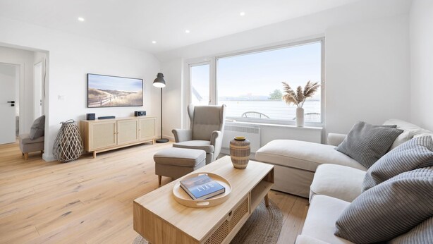 A modern living room with a sofa, armchair, and coffee table, a television, and a view of the sea through large windows.