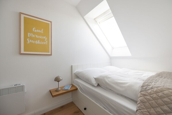 A bedroom with a sloped ceiling, a skylight, a bedside table with a lamp, and a framed quote on the wall.