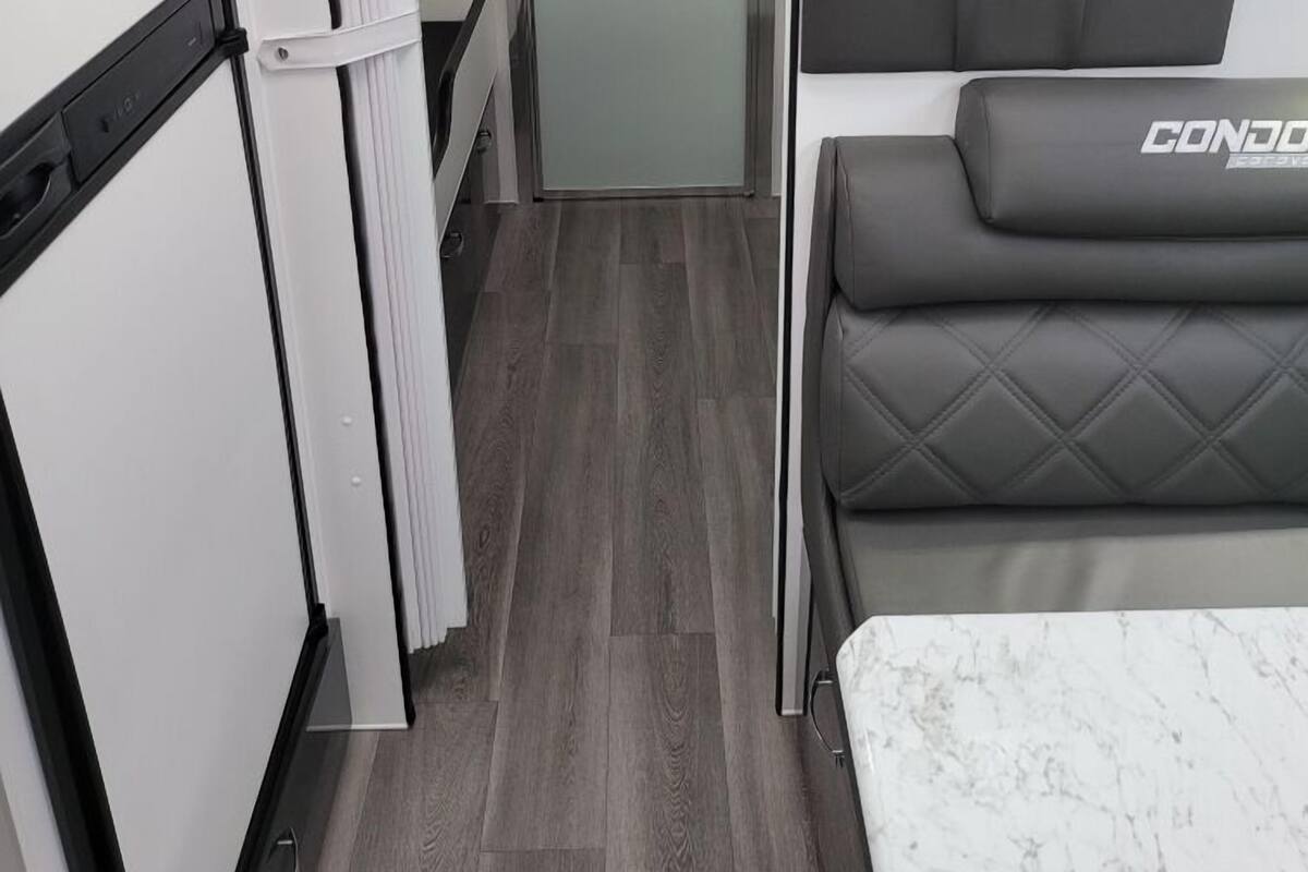 A compact, modern RV interior with a kitchenette, a sleeping area with a 'CONDO' branded bed, and a small bathroom.