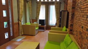A cozy living room with a brick wall, a wooden door, and a television. There are two green sofas and a wooden coffee table. - Kampung Turis Resort & Waterpark (Tegalwaru)