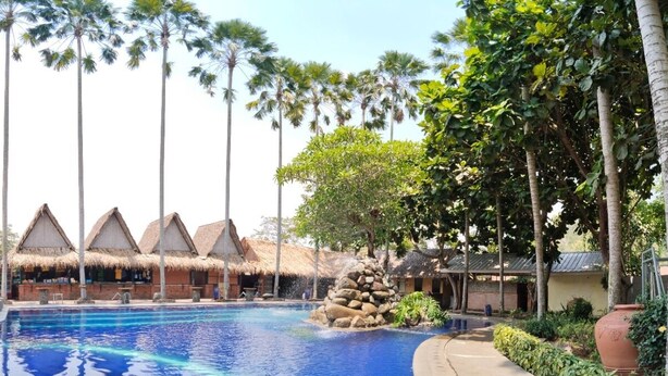 A pool area with thatched-roof structures, palm trees, and a clear blue pool. - Kampung Turis Resort & Waterpark (Tegalwaru)