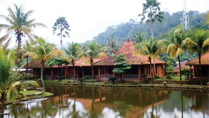 A traditional building with a tiled roof, surrounded by a calm water body and palm trees. - Kampung Turis Resort & Waterpark (Tegalwaru)
