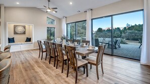 A spacious dining area with a large table and chairs, large windows offering a view of the outdoors, and a ceiling fan.