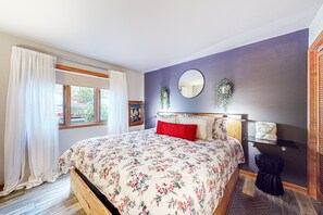 Quarto com colcha floral, espelho redondo, prateleira com toalhas e janela com cortinas.