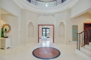 A spacious lobby with a central marble floor, a staircase on the right, and a view of the outside through an open door.