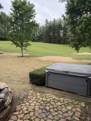 A stone patio with a hot tub, a small hedge, and a tree in a grassy field. - Indoor Pool + Hot Tub + Fire Pit + Game Room (Sevierville)