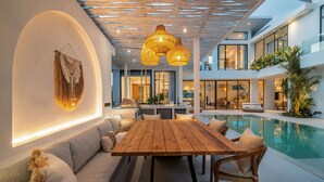 A modern indoor pool area with a wooden table and seating, surrounded by large windows and a ceiling with woven light fixtures.