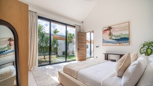A bedroom with a large bed, a sliding glass door to a balcony, a small table, and a painting on the wall.