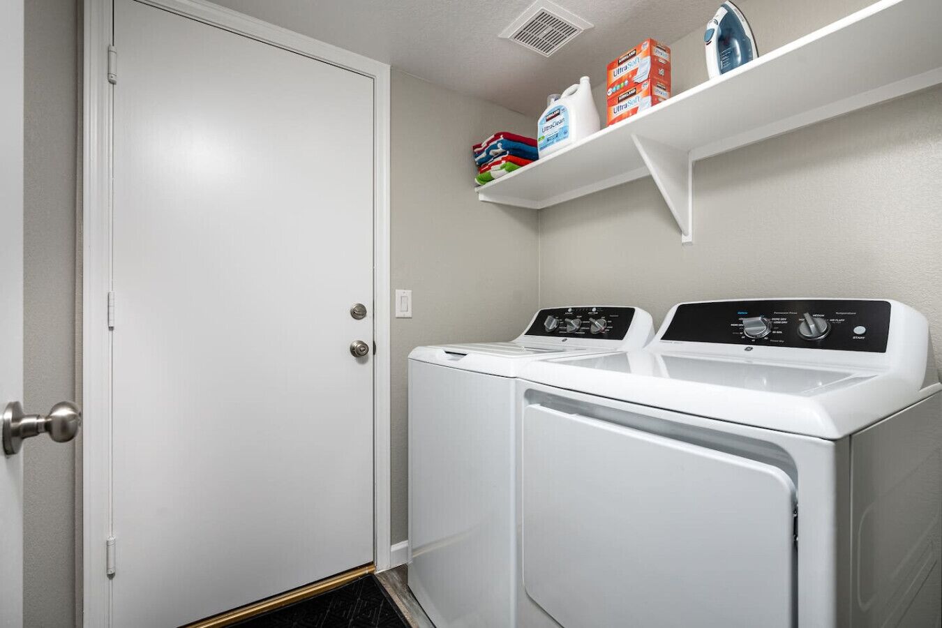 A laundry room with two white front-loading washers and dryers, a white door, and shelves above holding laundry supplies.