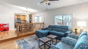 A spacious living room with a sectional sofa, ottomans, and a dining area with a wooden railing. - Spacious Family Home • Game Room • Pets Welcome (Orem)