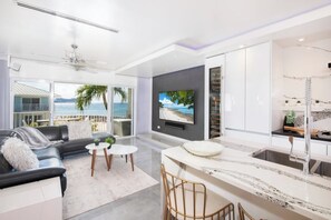 Uma sala de estar moderna com sofá, mesa de centro e TV, uma cozinha com bancada de mármore e pia, e uma varanda com vista para o oceano.