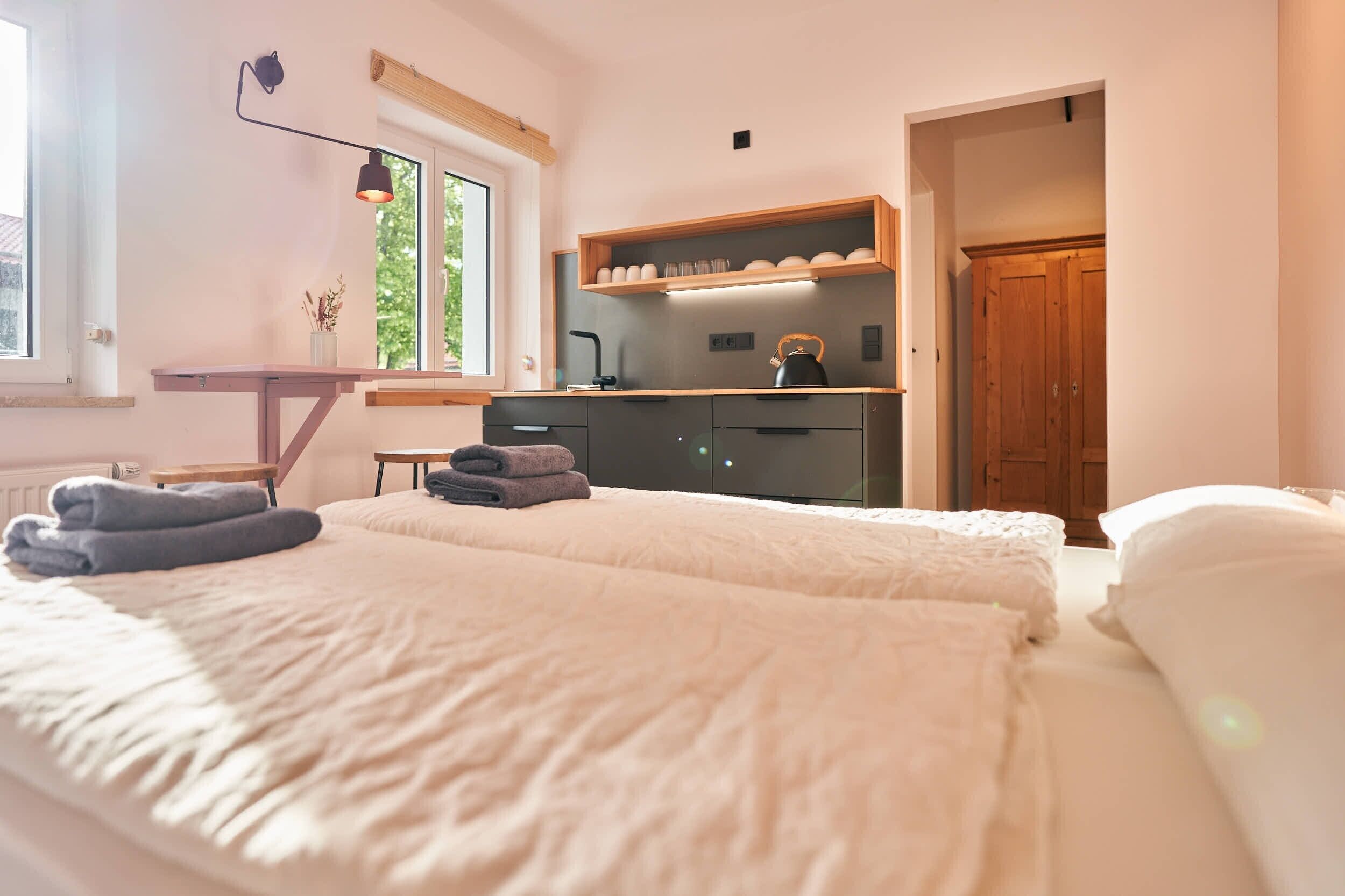 Un dormitorio con cama, escritorio, estante con tazas y vasos, y puerta que da a otra habitación.