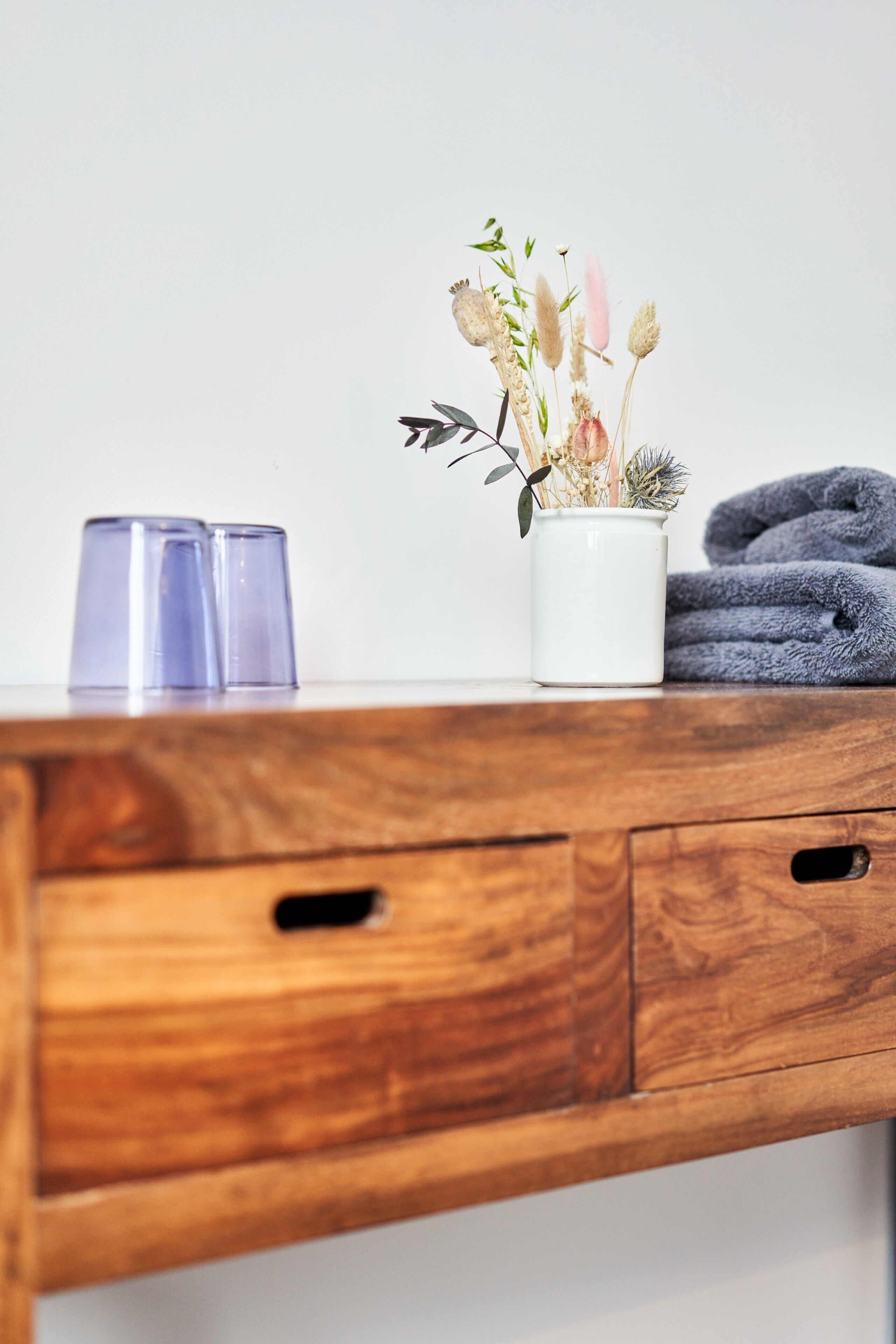 A wooden desk with a white vase of dried flowers, two clear glasses, and a folded gray towel.