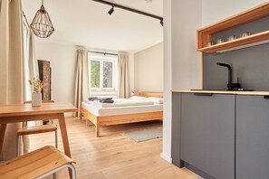 Un dormitorio con cama, un escritorio de madera, una silla, una ventana con cortinas, una lámpara colgante y una kitchenette con pileta y estantes.
