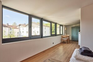 Una habitación con amplios ventanales, piso de madera, cama con almohadas, una mesa con una silla y un armario.
