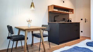 Ein gemütlicher Raum mit einem Holztisch, zwei Stühlen, einer kleinen Küchenzeile mit Wasserkocher und einem Bett mit blauer Decke.
