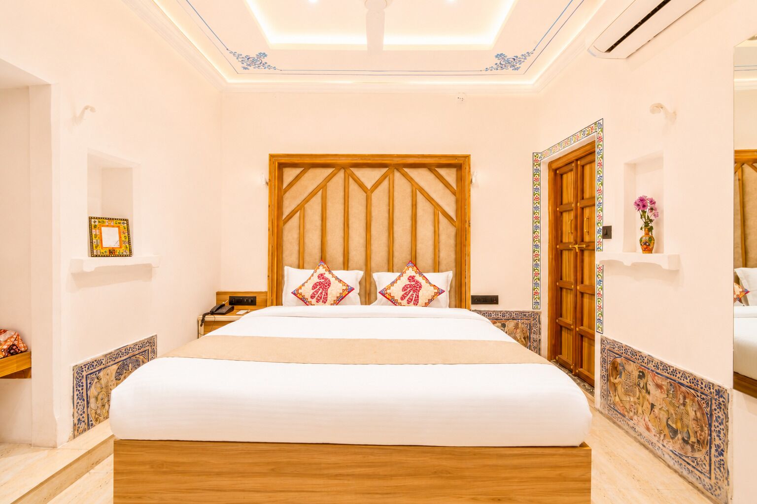 A hotel room with a large bed, wooden headboard, decorative pillows, a nightstand, a telephone, and a wall-mounted shelf with a vase.