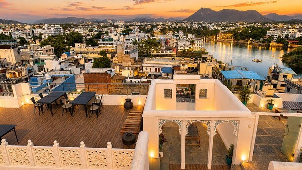 A rooftop terrace with tables and chairs, overlooking a cityscape and a river. - Amoraya — A Lake View Heritage Haveli (Old City Udaipur) (Udaipur)